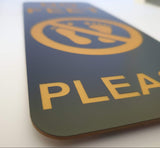 Close-up of a Black and gold acrylic sign with a logo and the word 'PLEASE'.