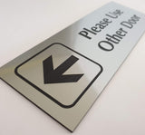 Silver acrylic sign with an arrow and black engraved  text 'Please Use Other Door' on a white background