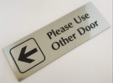 Silver acrylic sign with black engraved text 'Please Use Other Door' and an arrow