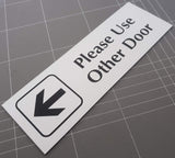 Rectangular White acrylic sign with Black engraved text 'Please Use Other Door' on a gray grid background
