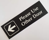 Black Rectangular acrylic sign with White engraved text 'Please Use Other Door' and an arrow on a white background