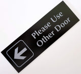 Black acrylic sign with Silver engraved text and an arrow pointing left on a white background
