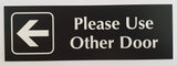 Black acrylic Sign with an arrow and white engraved text 'Please Use Other Door' on a white background