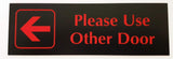 Black acrylic sign with red engraved arrow and text 'Please Use Other Door' on a white background