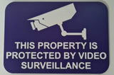 Sign with a camera icon and text 'This property is protected by video surveillance' on a purple background.