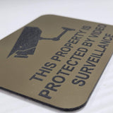 Gold acrylic sign with surveillance camera icon and text in Black background