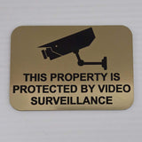 Gold acrylic sign with a security camera icon and text 'This property is protected by video surveillance' in Black.