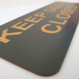 Black sign with gold text 'KEEP DOOR CLOSED' on a white background