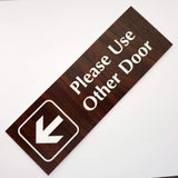 English Walnut acrylic sign with white engraved text 'Please Use Other Door' and an arrow on a white background