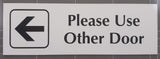 White acrylic Sign with an arrow pointing left and black engraved text 'Please Use Other Door' on a gray background