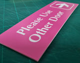 Pink acrylic sign with 'Please Use Other Door' white engraved text