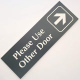 Rectangular smoke Grey acrylic sign with white engraved text 'Please Use Other Door' and an arrow on a white background