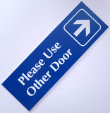 Blue acrylic sign with white engraved text and arrow on a light gray background