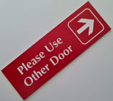 Red acrylic sign with white engraved text and arrow on a light gray background