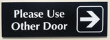 Black acrylic Sign with white engraved text 'Please Use Other Door' and an arrow on a black background