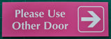 Pink acrylic sign with white engraved text 'Please Use Other Door' and an arrow on a green grid background